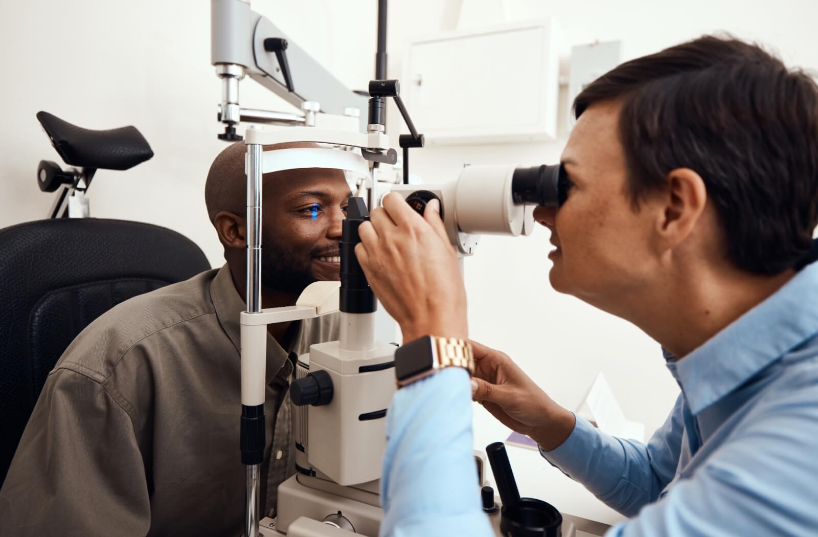 A patient sits in the examination chair at an optometrist's office. The optometrist sits across from the patient, examining the patient's eye through a tonometer.